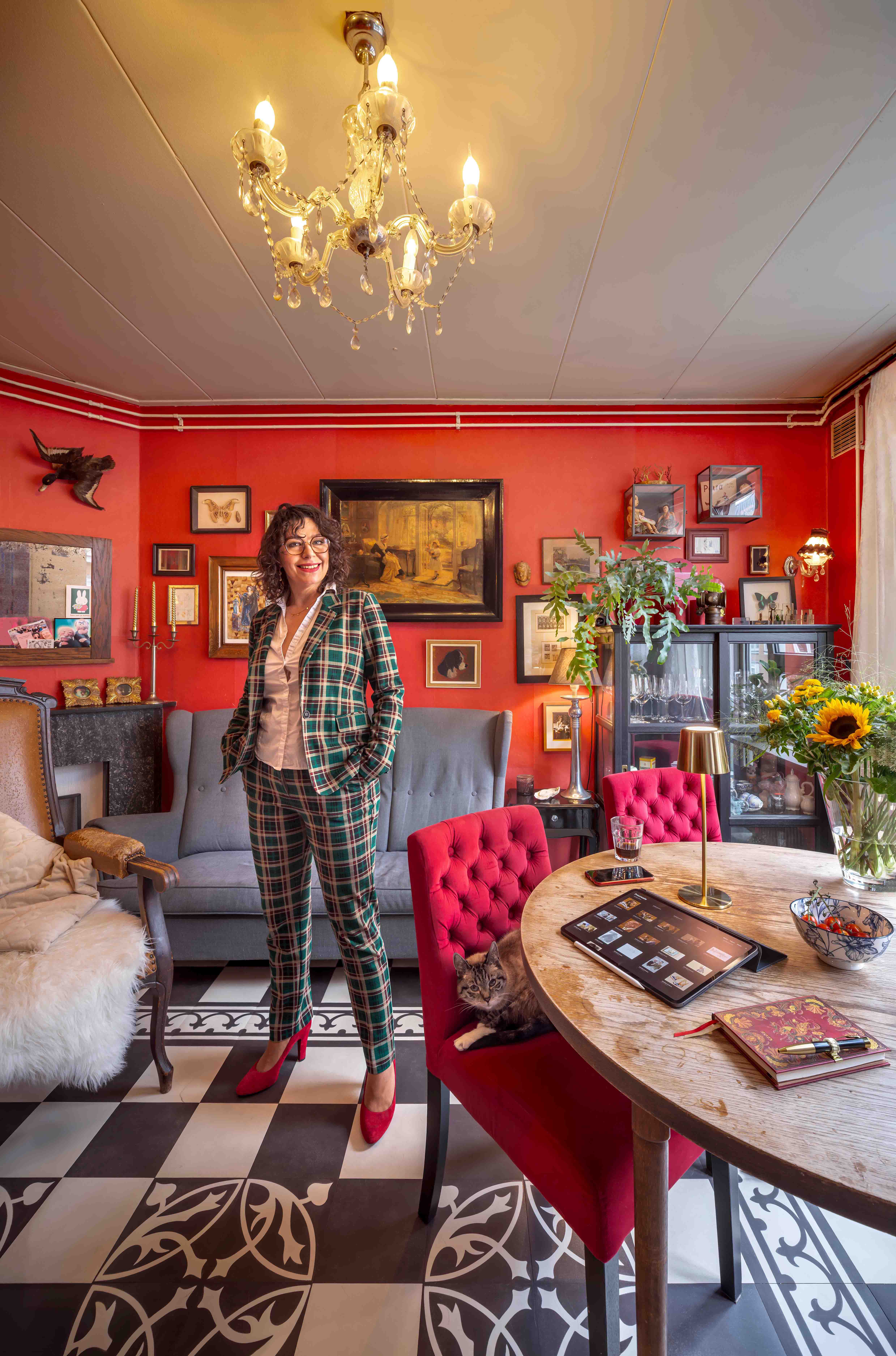 Petra Urban poseert in haar sfeervolle woonkamer met rode muren, vintage meubels en een kat op een rode fluwelen stoel.