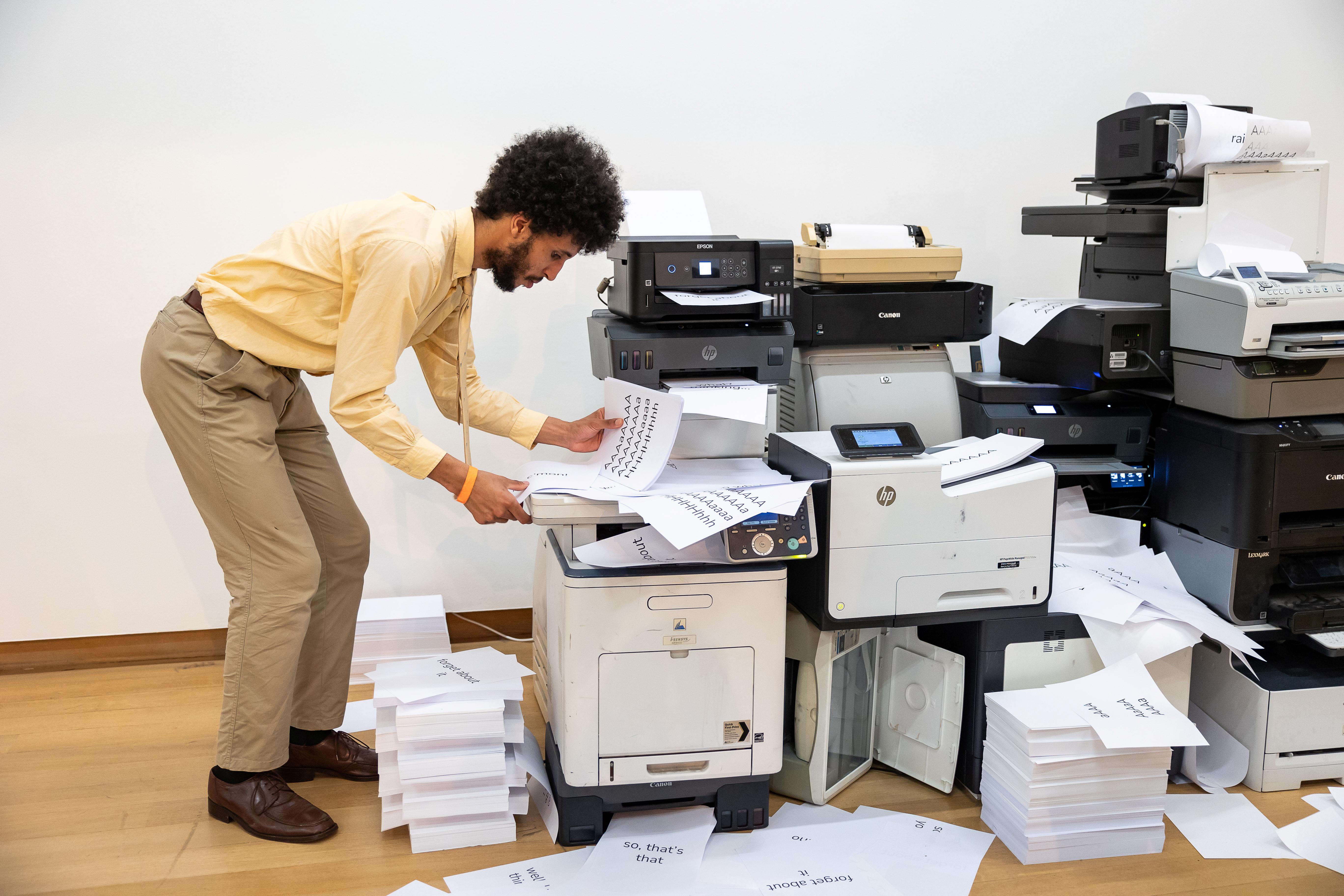Afbeelding van Rietveld Uncut in 2024. Er staan 10 printers op elkaar gestapeld met veel papier eromheen. Een jongen staat voorovergebogen ernaast en pakt 1 vel papier.